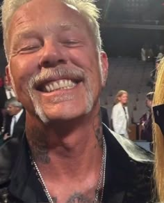 an older man with white hair and tattoos smiles at the camera while standing in front of a crowd