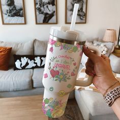 a woman is holding up a travel mug in her living room with pictures on the wall