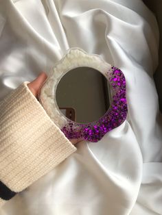 a hand holding a mirror with purple glitters on it and a white blanket in the background