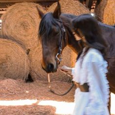 a woman walking next to a brown horse near hay bales in a barn area