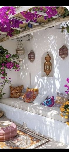 an outdoor seating area with purple flowers on the wall and potted plants hanging from the roof