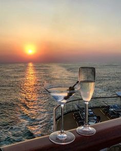 two wine glasses sitting on top of a wooden rail next to the ocean at sunset