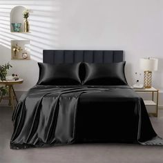 a bed with black sheets and pillows in a white room next to a lamp on a table