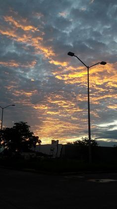 the sun is setting behind some street lights and trees in front of a cloudy sky