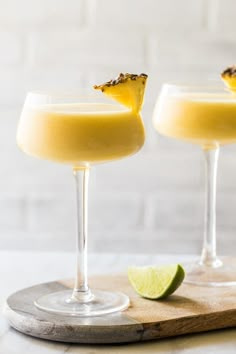 two glasses filled with drinks sitting on top of a cutting board next to a slice of lime