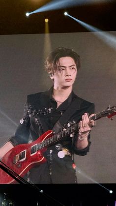 a man holding a red guitar while standing in front of a screen with lights on it