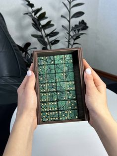 a person holding up a small box with green tiles on it and plants in the background