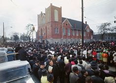 a large group of people standing in front of a building on a street with cars