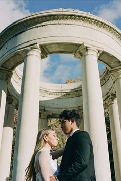 a man and woman standing next to each other in front of a white structure with columns