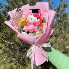 a hello kitty graduation bouquet is being held by someone's hand in front of some pine trees