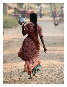 a woman walking down a dirt road carrying a metal bowl on her head and wearing a pink dress