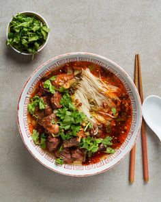 a bowl of soup with meat, noodles and greens next to chopsticks on a table