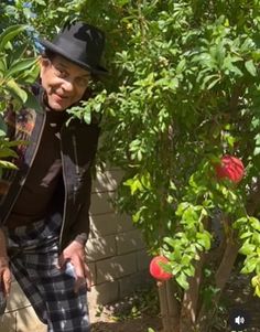 an older man wearing a black hat and plaid pants standing in front of a tree