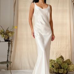 a woman in a long white dress standing next to a flower vase and curtained window