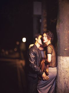 a man and woman standing next to each other near a tree on the street at night