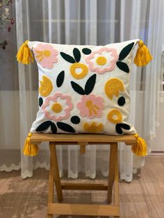 a white pillow with yellow and pink flowers on it sitting on a wooden stool in front of a window