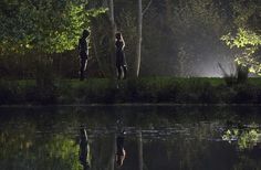 two people standing on the edge of a body of water in front of some trees