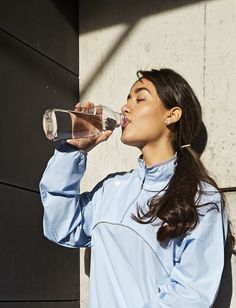 a woman drinking water from a bottle while standing next to a black wall with her eyes closed