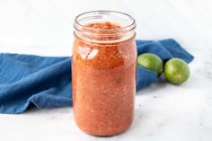 a jar filled with red sauce next to two limes on a blue towel and a white table cloth