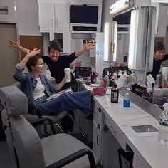 two people sitting in a hair salon chair with their arms up and one person standing