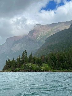 there is a boat on the water in front of some mountains and pine trees with clouds