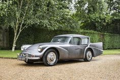 an old silver car parked in front of a hedged area next to a tree