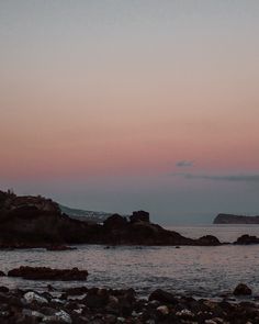 the sky is pink and purple as the sun sets over the ocean with rocks in the foreground