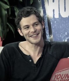 a young man sitting in front of a sign and smiling at the camera while wearing a black shirt