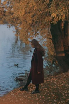 a woman in a red coat is standing by the water and ducks are swimming behind her