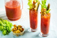 two glasses filled with red liquid and olives next to some other food on a table