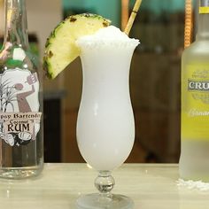 a white drink with a pineapple garnish on the rim and two bottles behind it