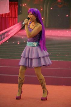 a woman with purple hair is singing into a microphone in front of some stairs and stage lights