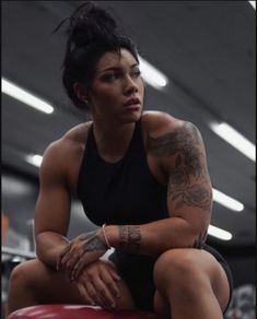 a woman with tattoos sitting on top of a red stool in a gym area,