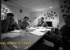 three people sitting at a table with papers and pens in front of them, one person holding his head