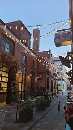 people sitting at tables on the sidewalk in front of some buildings with lights strung over them