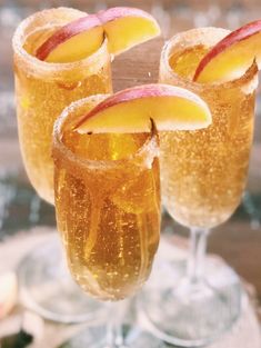 three glasses filled with apple cider on top of a wooden table next to an orange slice