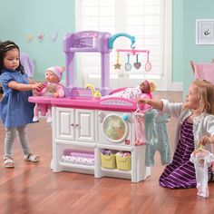 two girls playing with toys in a play kitchen and dining room area, while another girl watches from the other side