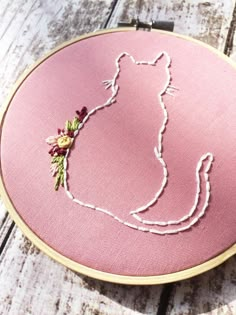 a cat with flowers on it's back in a pink embroidery hoop hanging from a wooden table