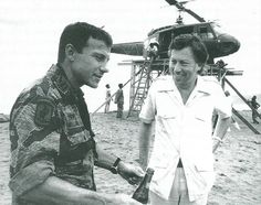 two men standing next to each other in front of a helicopter on top of a beach