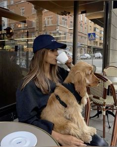 a woman sitting at an outdoor table drinking from a coffee cup while holding her dog