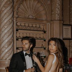 a man and woman standing next to each other in front of a shelf with wine glasses