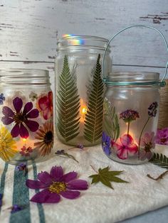 three glass jars with flowers and leaves in them sitting on a white towel next to some candles