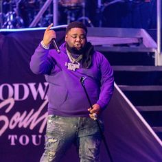 a man standing on top of a stage with a microphone in his hand and wearing a purple hoodie