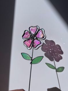 a hand holding a glass flower in front of a white wall with purple and green flowers