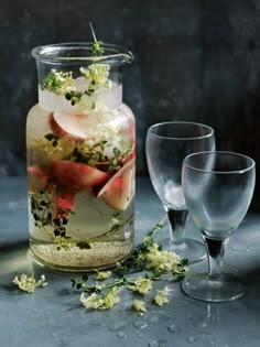 a jar filled with water and flowers next to two wine glasses on a gray surface