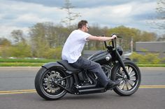 a man riding on the back of a black motorcycle down a street with trees in the background