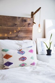 a white bed topped with pillows next to a wooden headboard and string lights on the wall