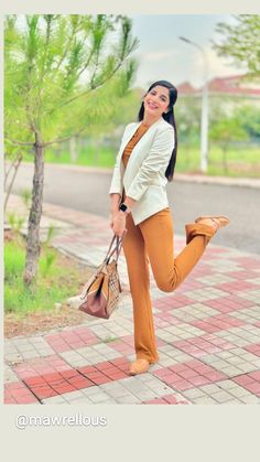 a woman in orange pants and a white blazer is posing on the sidewalk with her handbag