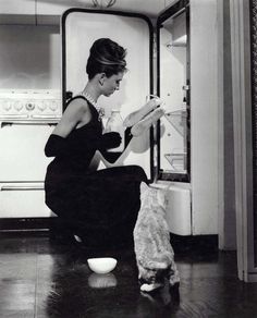 a woman sitting on the floor reading a book next to a cat in front of an open refrigerator