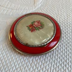 a small red and white compact with flowers on the lid sitting on a tablecloth